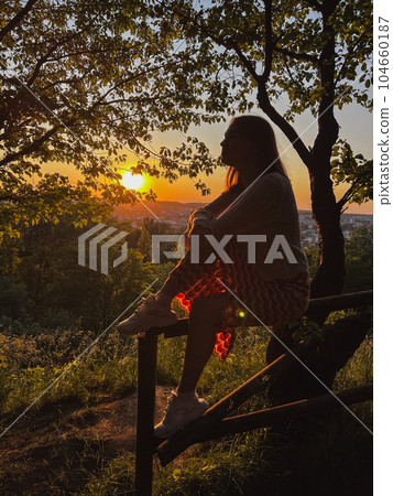 A woman sits on a fence overlooking the city and the sunset A woman sits on a fence overlooking the city and the sunset 104660187