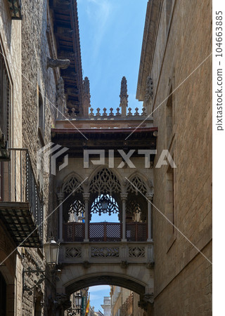 Bridge between buildings in Barri Gotic quarter of Barcelona, Spain. Pont del Bisbe at Dawn - A morning view of a neo-Gothic style stone bridge, "Pont del Bisbe" - Bishop's Bridge, over a narrow stone 104663885