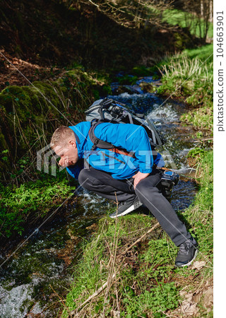 Hiker drinking stream water in mountain. Adult man drinking water from the mountain creek. Tourist wearing casual clothes and sunglasses making a sip of mountain river water from the palms of his 104663901