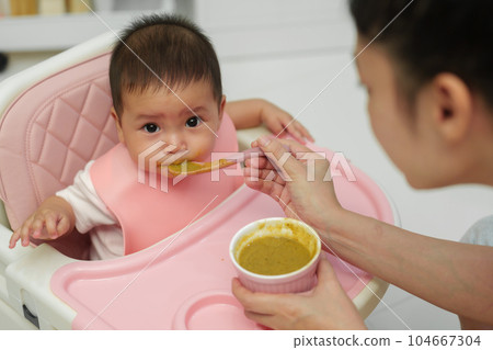 mother feeding food to her infant baby eating with spoon at home 104667304