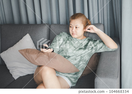 Asian housewife sit in sofa and holding remote control watching television with relaxing action. Hobby and free time life style concept. Asian housewife sit in sofa and holding remote control watching television with relaxing action. Hobby and free time life style concept. 104668334