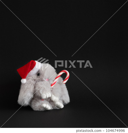Toy gray rabbit in Christmas Santa hat with candy cane on black background. 104674996