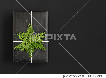 Black gift box with white ribbon, branches of thuja on black background with space for your text 104674999