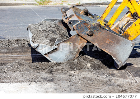 A fragment of the repair of an asphalt road section. The metal sliding bucket of a bulldozer cuts off the damaged part of the asphalt. 104677077