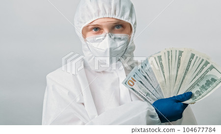 Healthcare salary. Doctor earnings. Female medical professional woman in white ppe blue gloves with dollar money fan isolated on grey background. 104677784