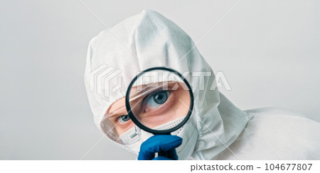 Medical virologist. Infection research. Female woman doctor technician lab specialist in white ppe hood protective equipment with magnifying glass isolated on grey background. Medical virologist. Infection research. Female woman doctor technician lab specialist in white ppe hood protective equipment with magnifying glass isolated on grey background. 104677807