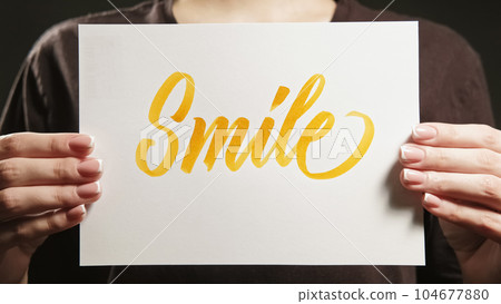 Smile sign. Positive motivation. Encouraging message. Unrecognizable woman supporting with optimistic support word text placard in hands. 104677880