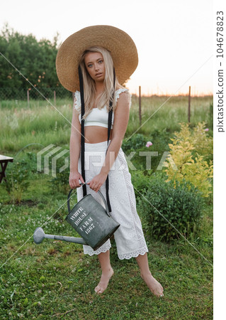 A blonde woman is watering flowers in the garden in the courtyard of a village house. A blonde woman is watering flowers in the garden in the courtyard of a village house. 104678823