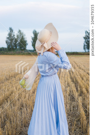A red-haired woman in a hat and a blue dress walks in a field with haystacks. 104679355