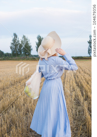A red-haired woman in a hat and a blue dress walks in a field with haystacks. 104679356