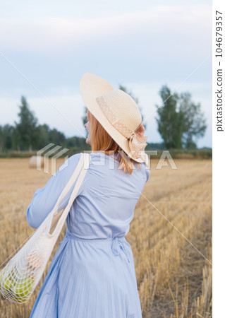 A red-haired woman in a hat and a blue dress walks in a field with haystacks. A red-haired woman in a hat and a blue dress walks in a field with haystacks. 104679357
