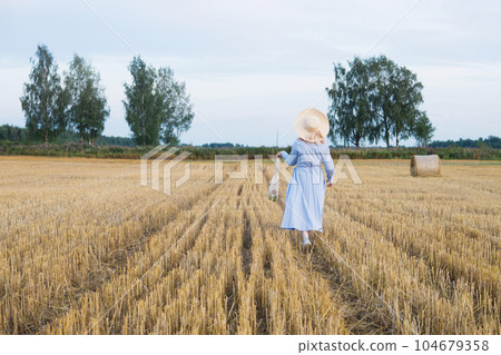 A red-haired woman in a hat and a blue dress walks in a field with haystacks. A red-haired woman in a hat and a blue dress walks in a field with haystacks. 104679358