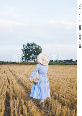 A red-haired woman in a hat and a blue dress walks in a field with haystacks. A red-haired woman in a hat and a blue dress walks in a field with haystacks. 104679359