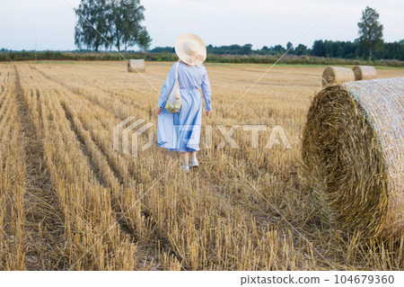 A red-haired woman in a hat and a blue dress walks in a field with haystacks. A red-haired woman in a hat and a blue dress walks in a field with haystacks. 104679360