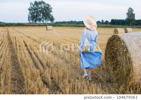 A red-haired woman in a hat and a blue dress walks in a field with haystacks. A red-haired woman in a hat and a blue dress walks in a field with haystacks. 104679361