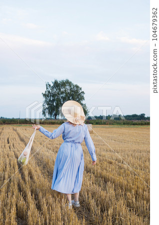 A red-haired woman in a hat and a blue dress walks in a field with haystacks. A red-haired woman in a hat and a blue dress walks in a field with haystacks. 104679362