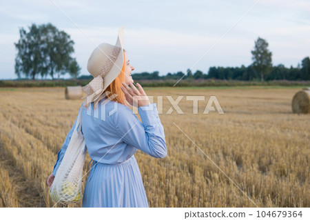 A red-haired woman in a hat and a blue dress walks in a field with haystacks. A red-haired woman in a hat and a blue dress walks in a field with haystacks. 104679364