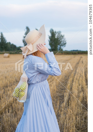 A red-haired woman in a hat and a blue dress walks in a field with haystacks. A red-haired woman in a hat and a blue dress walks in a field with haystacks. 104679365