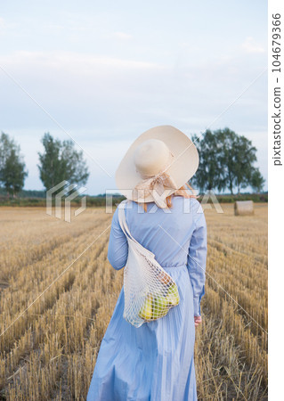 A red-haired woman in a hat and a blue dress walks in a field with haystacks. A red-haired woman in a hat and a blue dress walks in a field with haystacks. 104679366