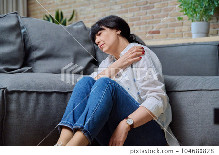 Mature woman suffering from pain in her shoulder, sitting at home on floor Mature woman suffering from pain in her shoulder, sitting at home on floor 104680828