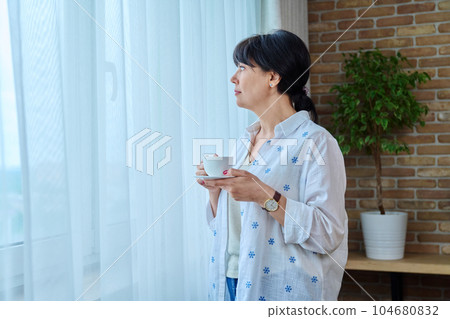Calm beautiful mature woman with cup of coffee at home near window 104680832