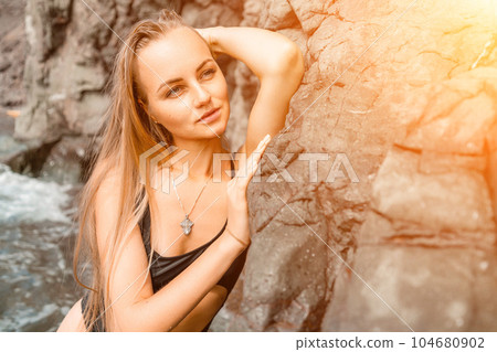 Woman swimsuit sea. Attractive blonde woman in a black swimsuit enjoying the sea air on the seashore around the rocks. Travel and vacation concept. 104680902