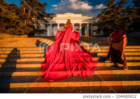 Sunrise red dress. A woman in a long red dress against the backdrop of sunrise, bright golden light of the sun's rays. The concept of femininity, harmony. 104680922