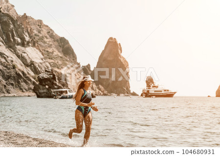 Woman travel summer sea. A happy tourist in a blue bikini enjoying the scenic view of the sea and volcanic mountains while taking pictures to capture the memories of her travel adventure. 104680931