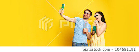 Summer vacation, Young couple, man and woman in sunglasses drinking lemonade and taking selfie against yellow studio background Summer vacation, Young couple, man and woman in sunglasses drinking lemonade and taking selfie against yellow studio background 104681191