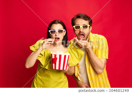 Man and woman emotionally watching movie, eating popcorn against red studio background. Shocked faces Man and woman emotionally watching movie, eating popcorn against red studio background. Shocked faces 104681249