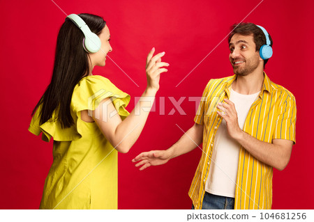 Positive young man and woman in bright yellow clothes listening to music in headphones and dancing against red studio background Positive young man and woman in bright yellow clothes listening to music in headphones and dancing against red studio background 104681256
