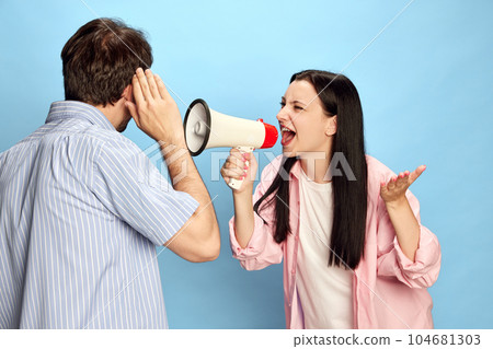 Woman shouting in megaphone to man against blue studio background. Emotional conversation and misunderstanding 104681303