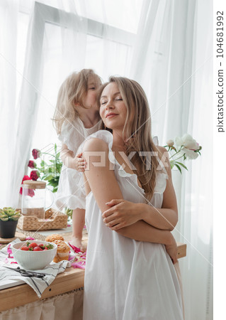 A little blonde girl with her mom on a kitchen countertop decorated with peonies. The concept of the relationship between mother and daughter. Spring atmosphere. A little blonde girl with her mom on a kitchen countertop decorated with peonies. The concept of the relationship between mother and daughter. Spring atmosphere. 104681992