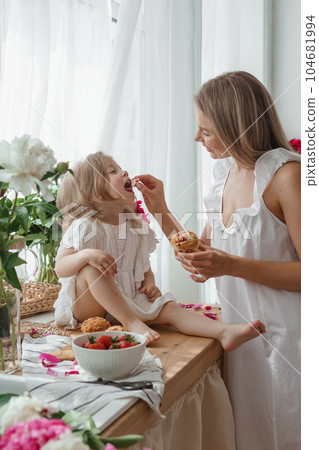 A little blonde girl with her mom on a kitchen countertop decorated with peonies. The concept of the relationship between mother and daughter. Spring atmosphere. A little blonde girl with her mom on a kitchen countertop decorated with peonies. The concept of the relationship between mother and daughter. Spring atmosphere. 104681994