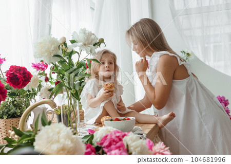 A little blonde girl with her mom on a kitchen countertop decorated with peonies. The concept of the relationship between mother and daughter. Spring atmosphere. A little blonde girl with her mom on a kitchen countertop decorated with peonies. The concept of the relationship between mother and daughter. Spring atmosphere. 104681996