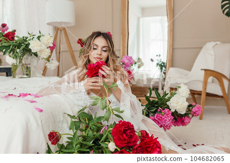 A beautiful girl in a white nightgown at home, surrounded by spring flowers on a large bed. A room decorated with bouquets of peonies 104682065