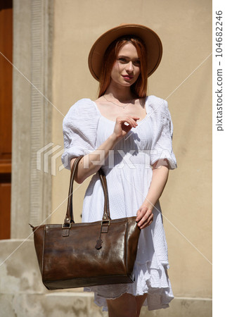 young beautiful redhair girl with red lipstick in a big hat and white summer dress with brown handbag posing on the streets 104682246
