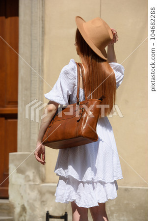 young beautiful redhair girl with red lipstick in her big hat and white summer dress with light brown handbag posing on the streets 104682288