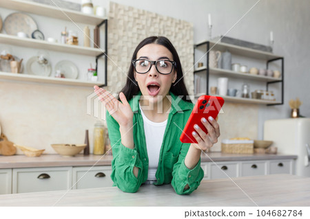 Shocked and surprised young woman looking at camera. Sitting in the kitchen at home, holding the phone and spreading his hands. 104682784