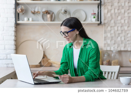 Beautiful young Muslim woman in hijab wearing headphones sitting in kitchen at home at table and using laptop. She listens to music, is satisfied, raises her hands up, dances along. 104682789