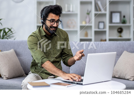 Young man working from home remotely, idus using headset phone and laptop for video call and remote communication with clients, businessman sitting at home in living room on sofa. Young man working from home remotely, idus using headset phone and laptop for video call and remote communication with clients, businessman sitting at home in living room on sofa. 104685284