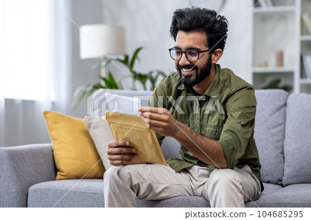 Joyful hispanic received letter mail notification man sitting at home in living room on sofa holding envelope smiling reading. Joyful hispanic received letter mail notification man sitting at home in living room on sofa holding envelope smiling reading. 104685295