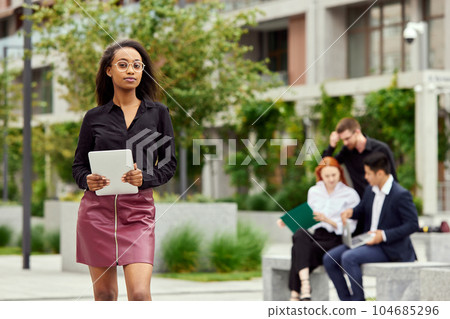 Portrait of concentrated young businesswoman in glasses standing with tablet on street. Blurred employees on background 104685296