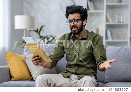 Upset and sad man at home received notification mail envelope with bad news, hindu man sitting on sofa in living room depressed reading and holding envelope with message in hands. Upset and sad man at home received notification mail envelope with bad news, hindu man sitting on sofa in living room depressed reading and holding envelope with message in hands. 104685297