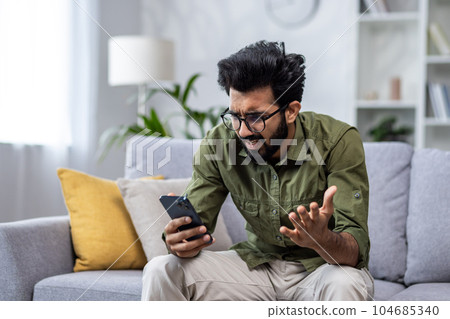 Upset and sad man sitting on sofa at home in living room, hindu man reading bad news online from phone, depressed and sad man. Upset and sad man sitting on sofa at home in living room, hindu man reading bad news online from phone, depressed and sad man. 104685340