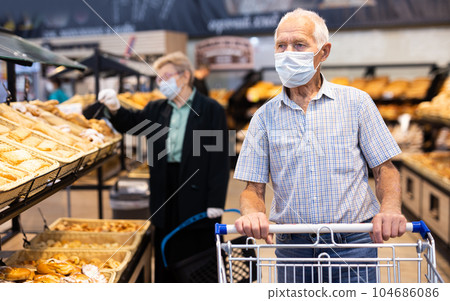mature european man wearing mask and gloves with covid protection chooses buns and bread in supermarket bakery 104686086