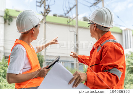 Back view of 2 male engineers or foremen in orange jumpsuit, safety vest and white hardhat, one holding a computer tablet and the other holding a building blueprint, working together pointing out the Back view of 2 male engineers or foremen in orange jumpsuit, safety vest and white hardhat, one holding a computer tablet and the other holding a building blueprint, working together pointing out the 104686593