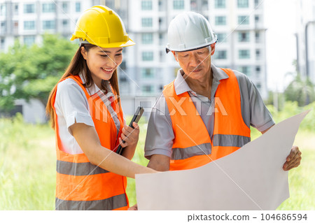 Serious male and happy female engineers looking at the construction blueprint together at an outdoor site with blurred buildings in background 104686594