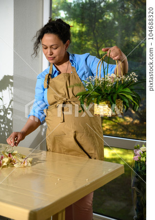 Beautiful pregnant woman in beige apron, arranging flower bouquet in wicker basket, working in floral design studio 104688320