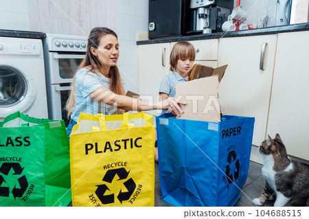 Mother is teaching kid how to recycle help the boy aware environmental importance - mom educates son sort garbage into different bins on kitchen. Family sorting garbage at home. Concept of recycling. Mother is teaching kid how to recycle help the boy aware environmental importance - mom educates son sort garbage into different bins on kitchen. Family sorting garbage at home. Concept of recycling. 104688515